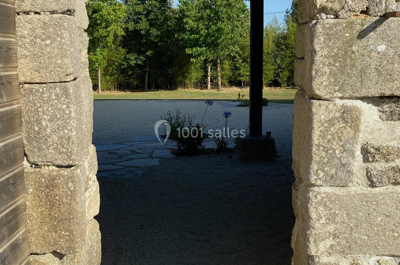 Vue à travers une porte en pierre donnant sur une cour en gravier avec des arbres et des fleurs en arrière-plan.
