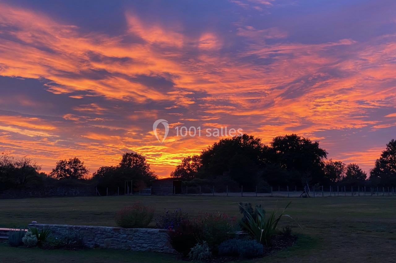 Ciel vibrant aux teintes orange et violette au coucher du soleil, avec des arbres et un jardin en premier plan.