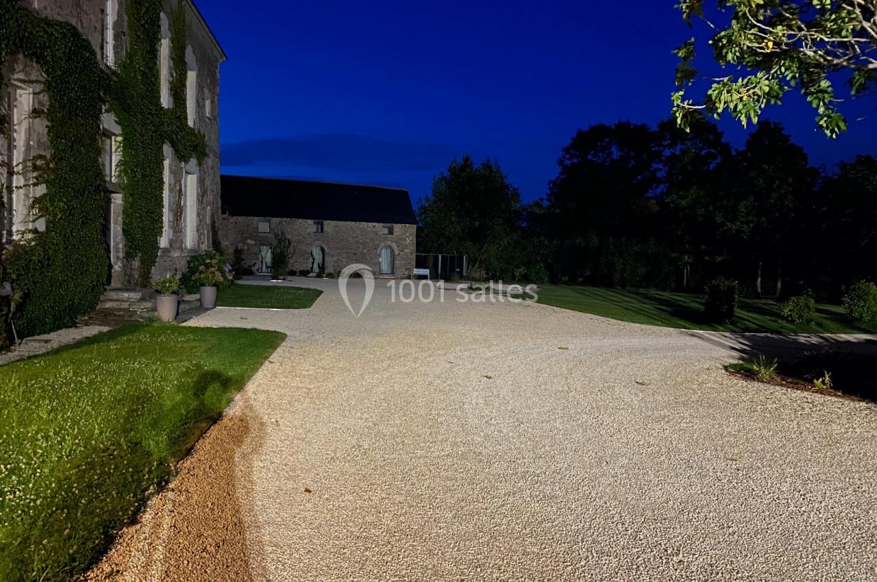 Allée en gravier éclairée la nuit, bordée de pelouse et d'un bâtiment en pierre recouvert de végétation.