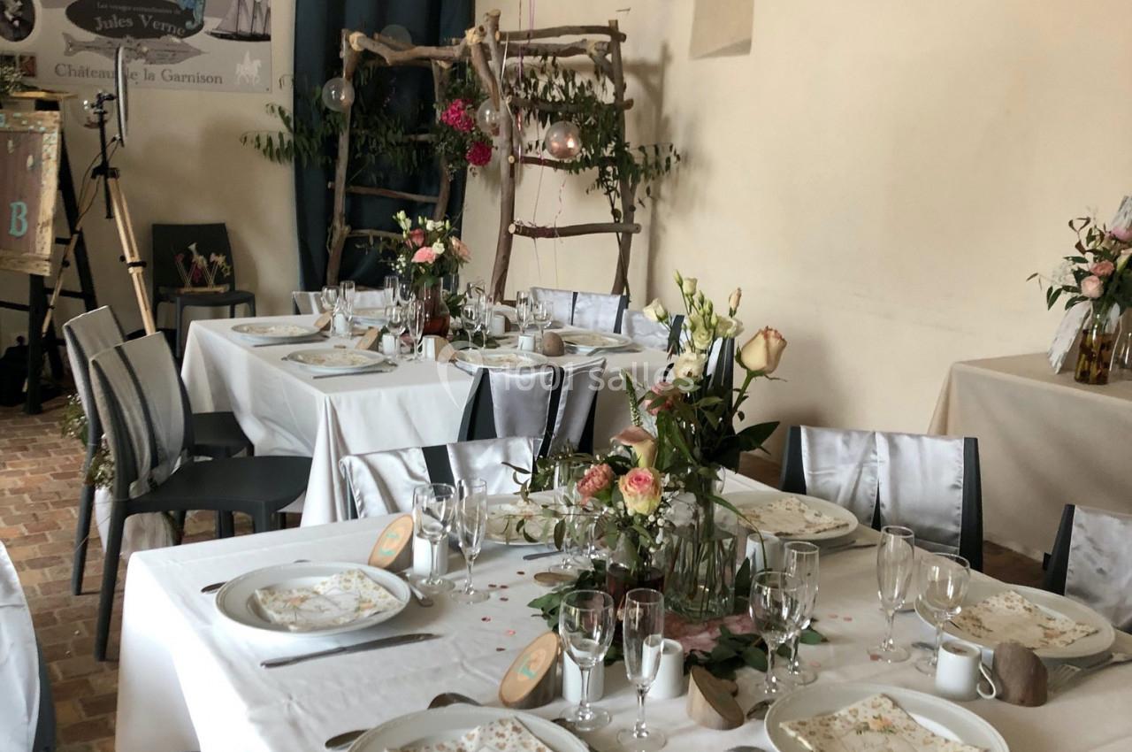 Salle décorée pour un repas festif, avec tables dressées, nappes blanches, fleurs et ballons roses en arrière-plan.