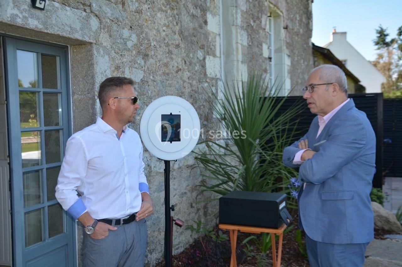Deux hommes discutent à l'extérieur près d'un mur en pierre, avec un dispositif circulaire et un haut-parleur à proximité.