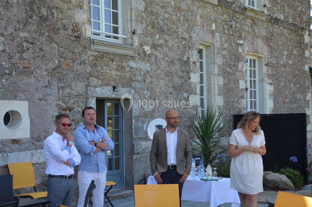 Quatre personnes debout devant un bâtiment en pierre, avec des chaises et une table dressée à proximité.