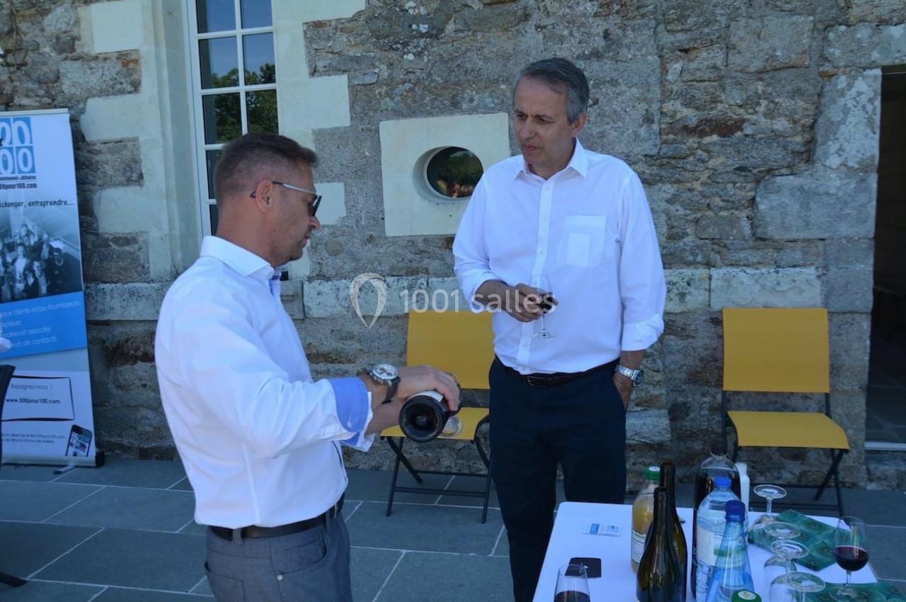 Deux hommes en chemise discutent autour d'une table avec des bouteilles et verres, devant un mur en pierre.