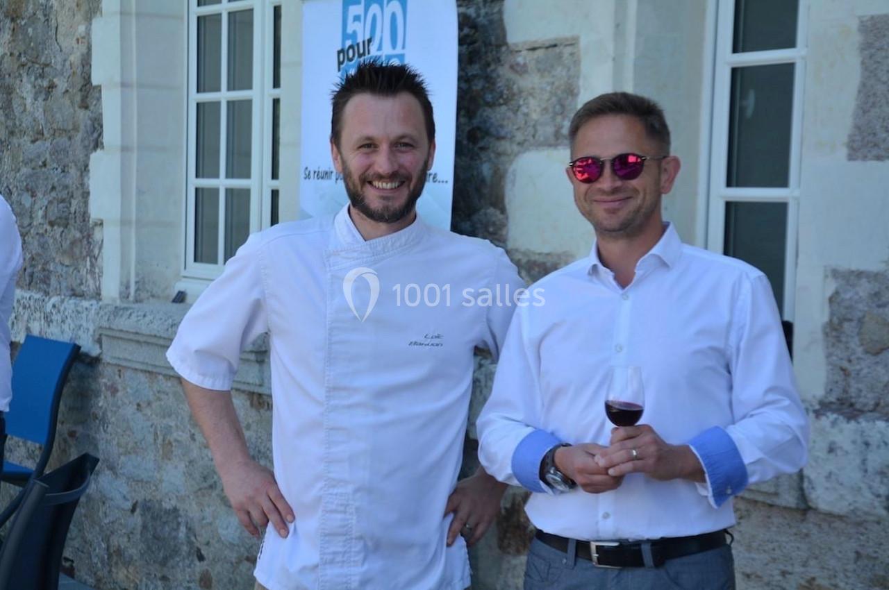 Deux hommes souriants, l'un en tenue de chef, l'autre en chemise blanche, tenant un verre de vin, devant un bâtiment en…