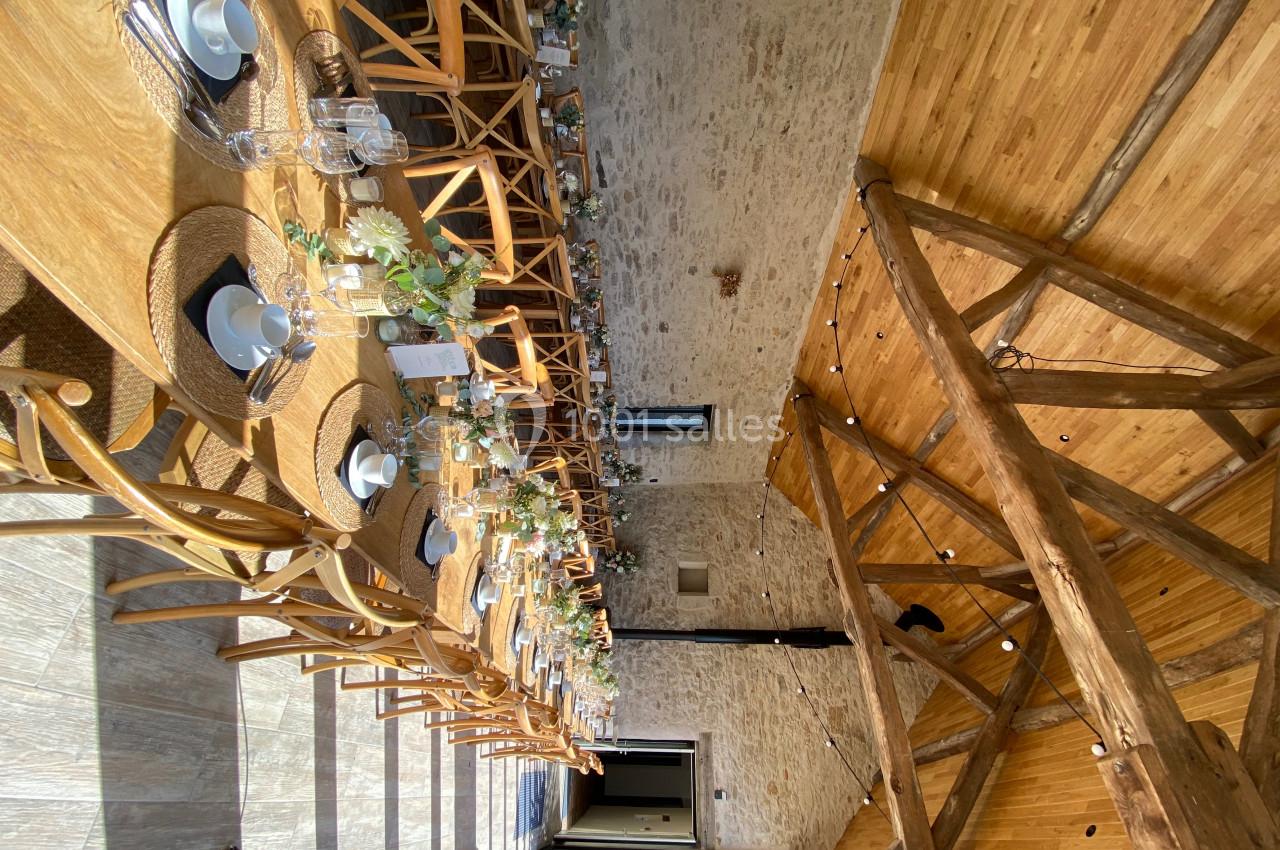 Salle en pierre avec de grandes tables en bois décorées de fleurs et de vaisselle, éclairée par une lumière naturelle.