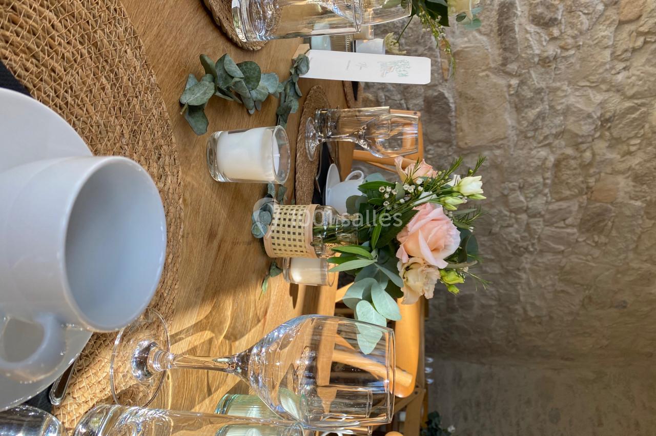 Table décorée avec des fleurs, bougies et vaisselle, sur une nappe en jute, dans une salle aux murs en pierre.