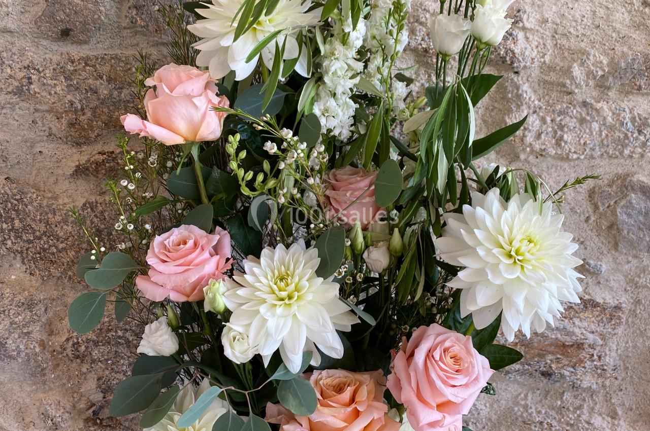 Arrangement floral avec roses roses, dahlias blancs et feuillage vert dans un vase rose, posé devant un mur en pierre.