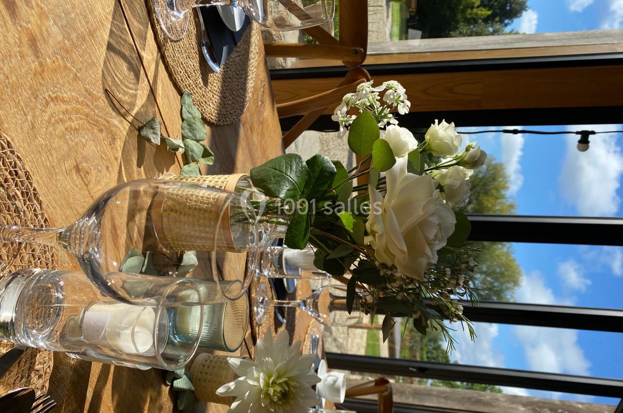 Table en bois dressée avec des verres, des bougies et un bouquet de fleurs blanches, près d'une grande baie vitrée.