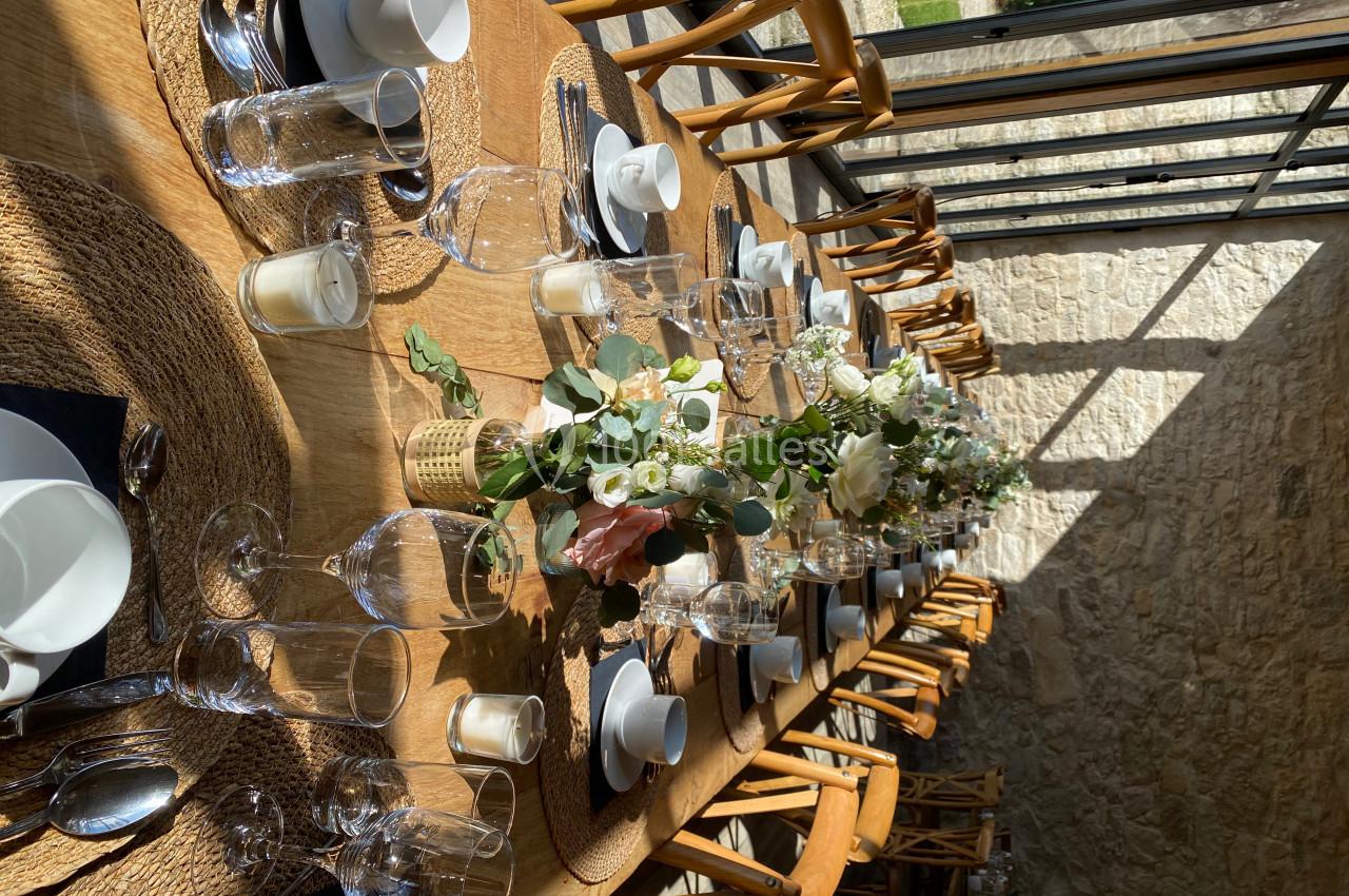 Table en bois dressée pour un repas, avec vaisselle élégante, verres, bougies et compositions florales au centre.
