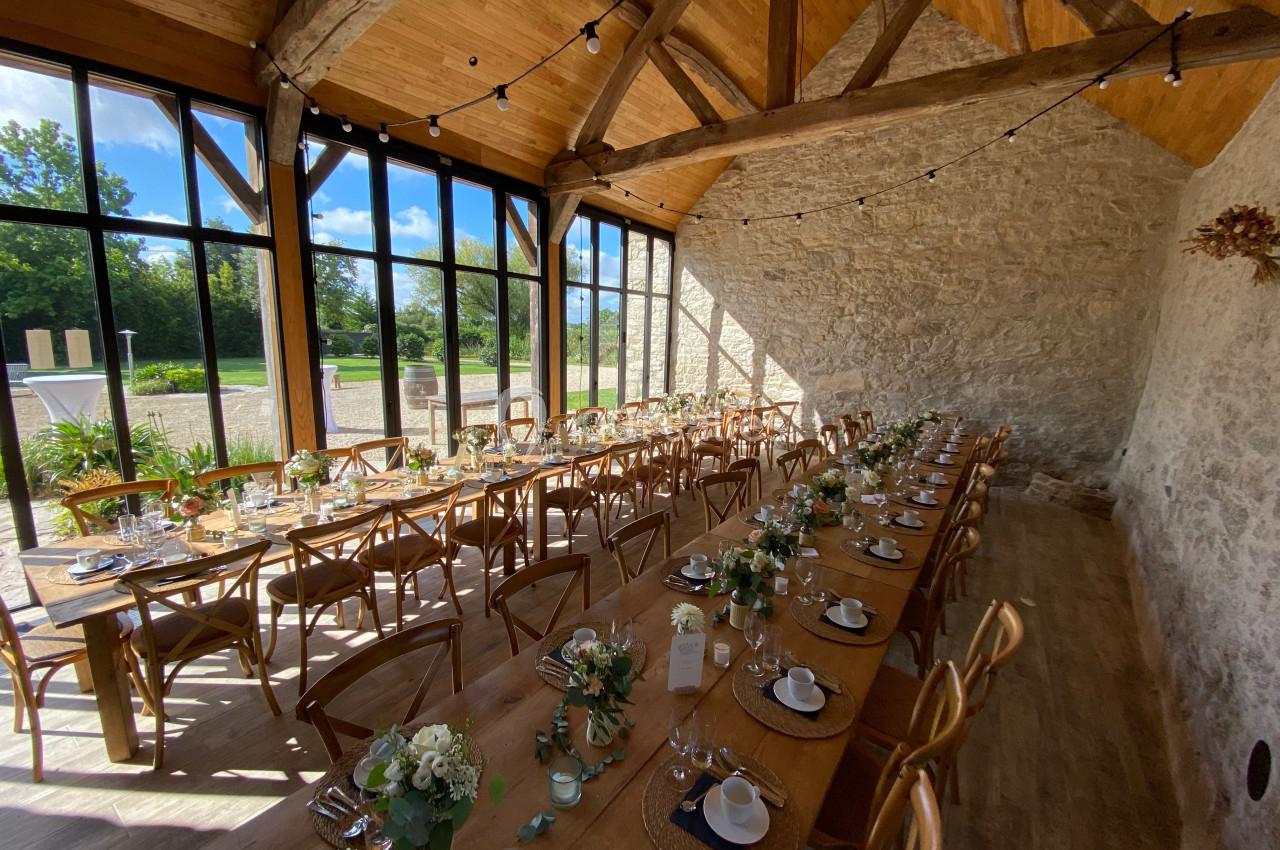 Grande salle lumineuse avec tables en bois décorées de fleurs, chaises assorties et poutres apparentes sous un plafond haut.