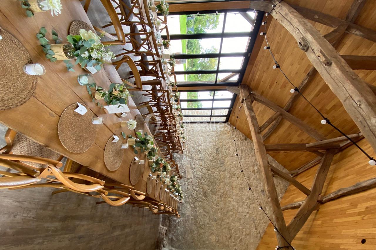 Grande table en bois décorée de fleurs et de sets en paille dans une salle avec poutres apparentes et grandes baies vitrées.