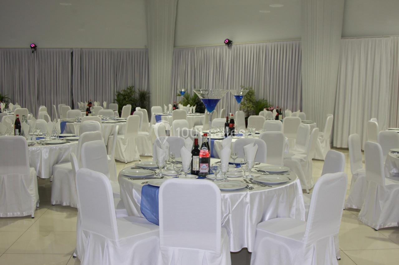 Salle de réception décorée avec des tables rondes dressées, nappes blanches et centres de table en verre.