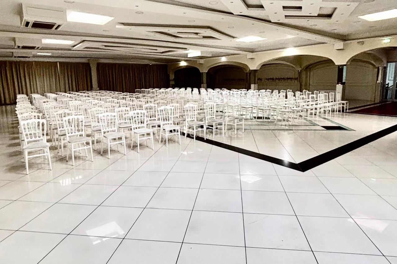 Salle de réception lumineuse avec des rangées de chaises blanches alignées sur un sol en carrelage clair.