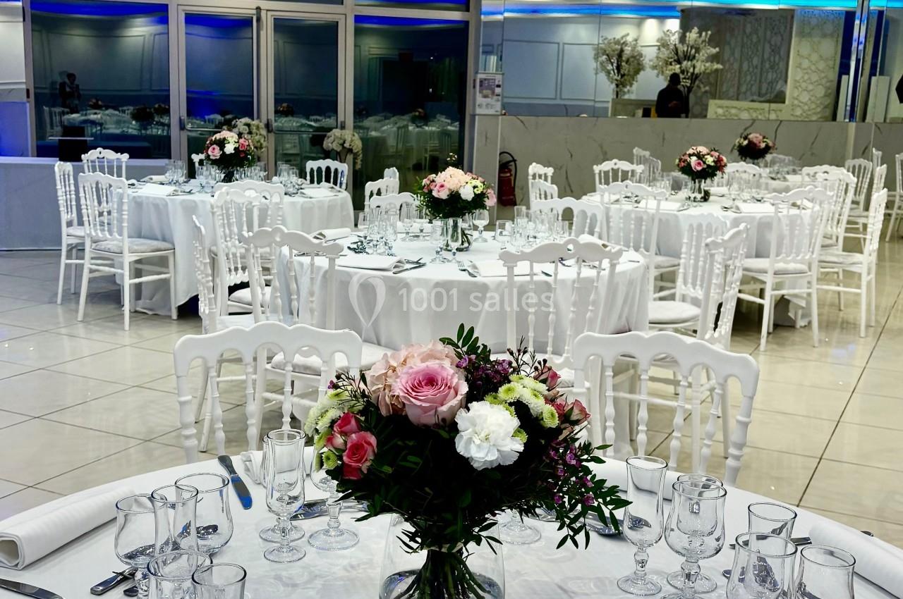 Salle de réception élégante avec tables rondes dressées, décorées de bouquets de fleurs et éclairage moderne.