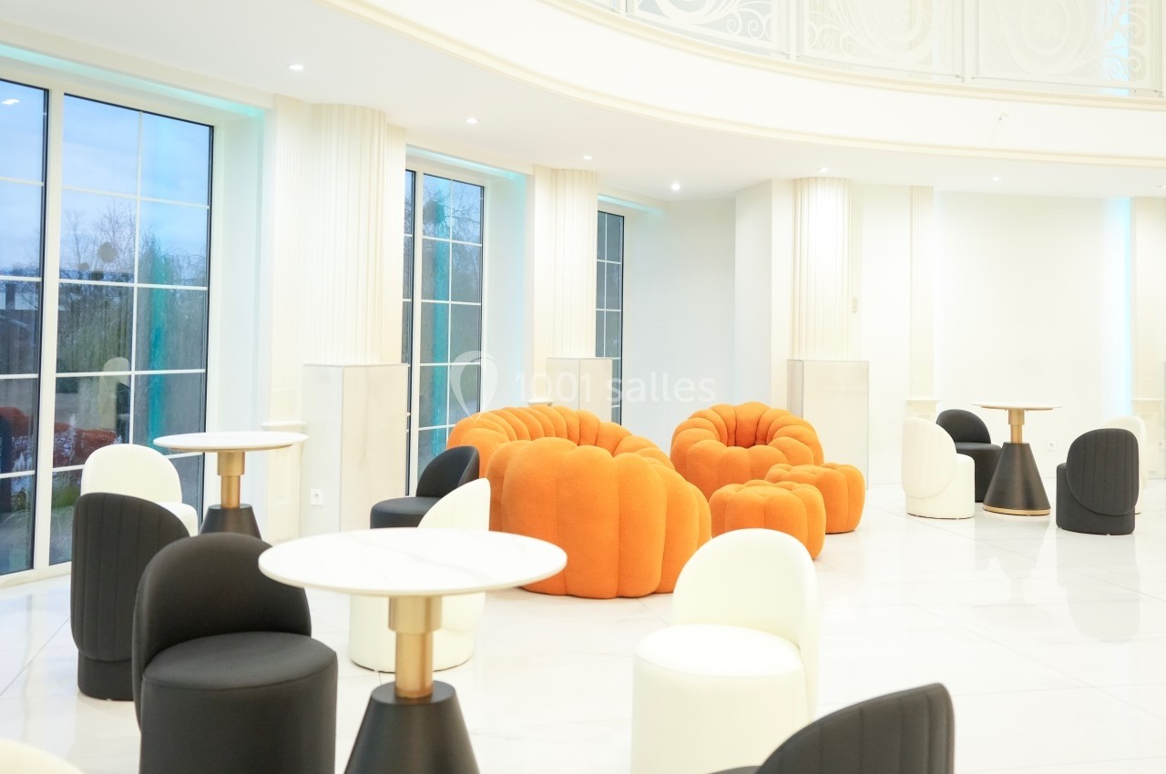 Espace lumineux avec fauteuils orange et noirs, tables rondes et grandes fenêtres donnant sur l'extérieur.