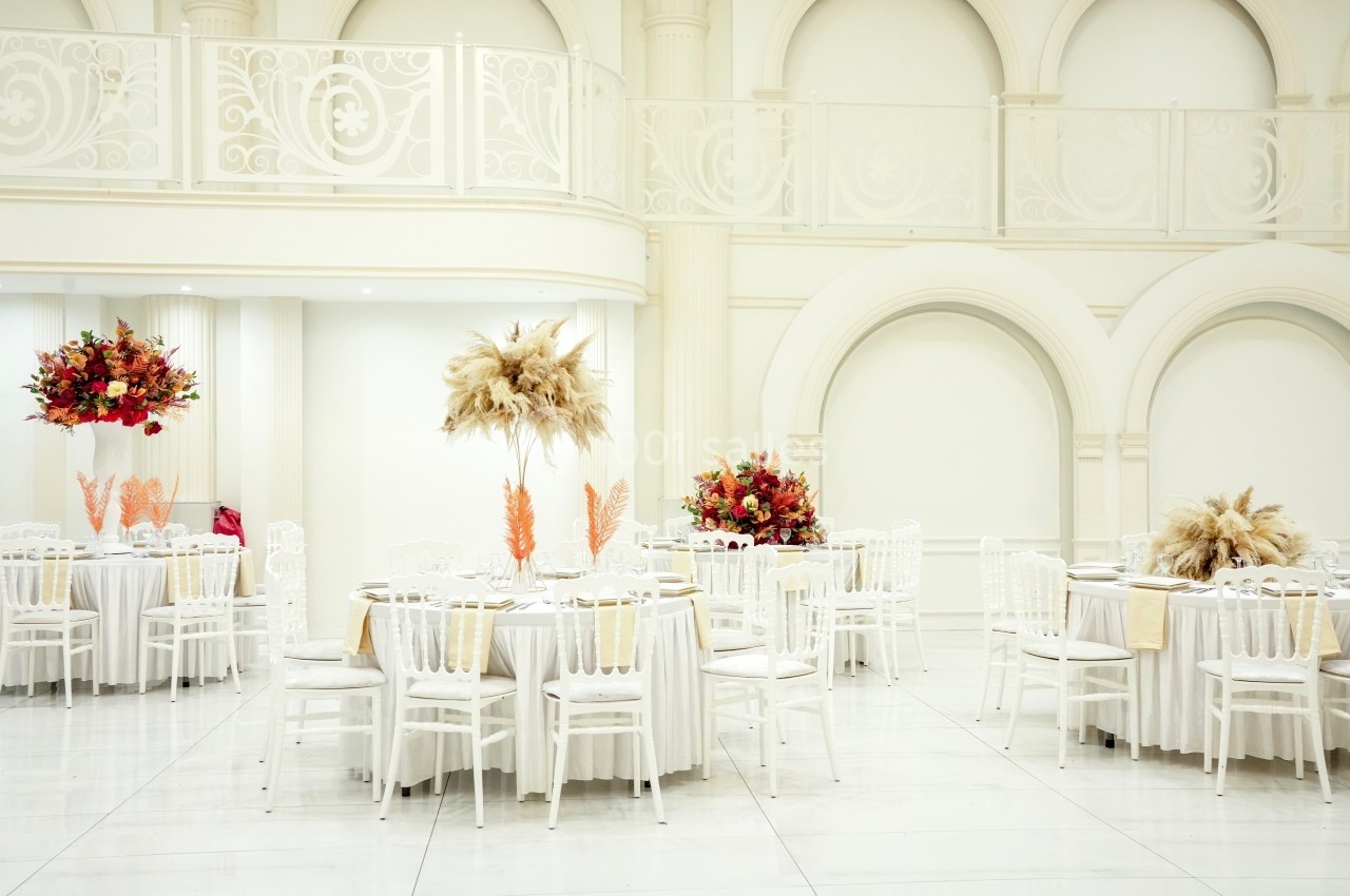 Salle de réception lumineuse avec tables rondes décorées de fleurs colorées et de pampas, entourées de chaises blanches.