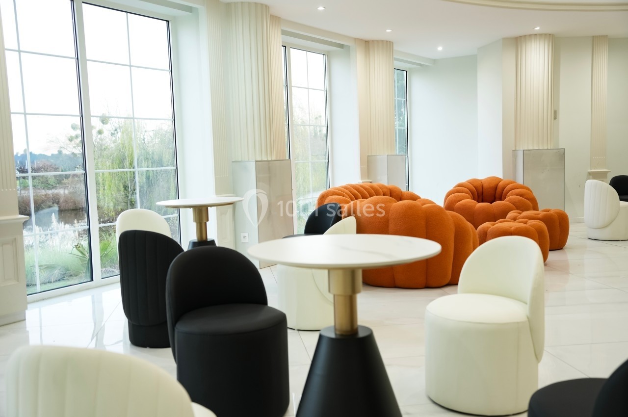 Salle lumineuse avec fauteuils design orange et noirs, tables blanches et grandes baies vitrées donnant sur un jardin.