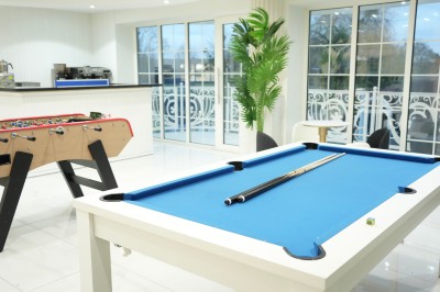 Table de billard bleu avec une queue et une bille, située dans une pièce lumineuse avec un baby-foot et de grandes baies…