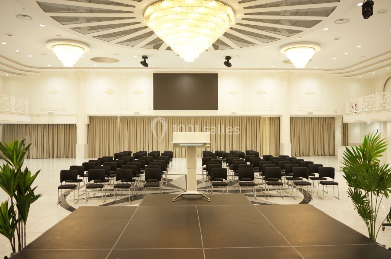 Salle de conférence lumineuse avec un podium, des rangées de chaises noires et un grand écran en arrière-plan.
