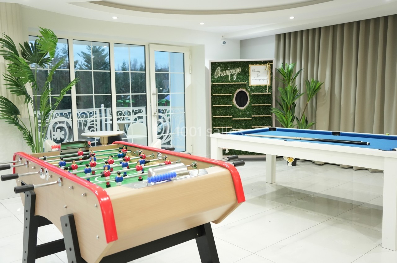 Salle de loisirs lumineuse avec baby-foot, table de billard, plantes et grande baie vitrée donnant sur l'extérieur.