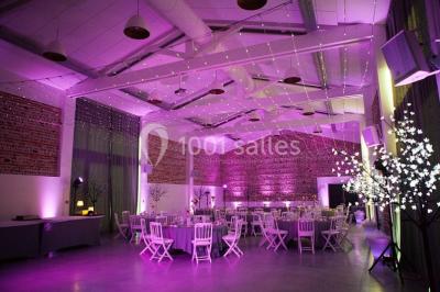 Salle de réception décorée pour un mariage, avec tables rondes, nappes blanches et guirlandes lumineuses au plafond.