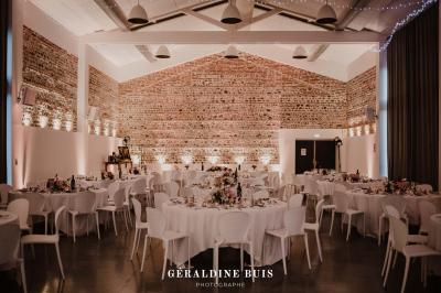 Salle de réception décorée pour un mariage, avec tables rondes, nappes blanches et guirlandes lumineuses au plafond.