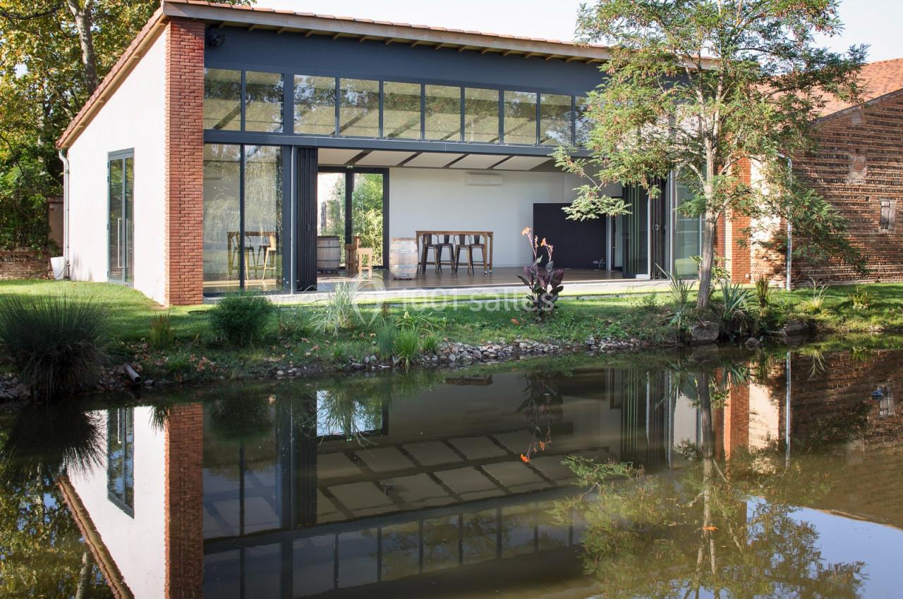 Maison moderne avec grandes baies vitrées, entourée de verdure et reflétée dans un étang calme.