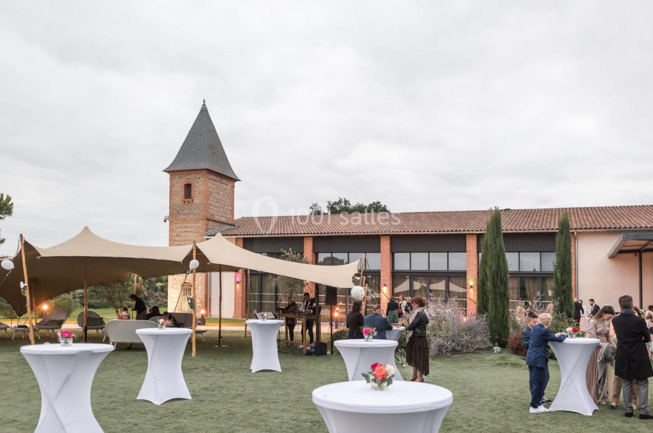 Jardin avec tables hautes décorées, invités rassemblés près d'une tente et d'un bâtiment en briques sous un ciel nuageux.