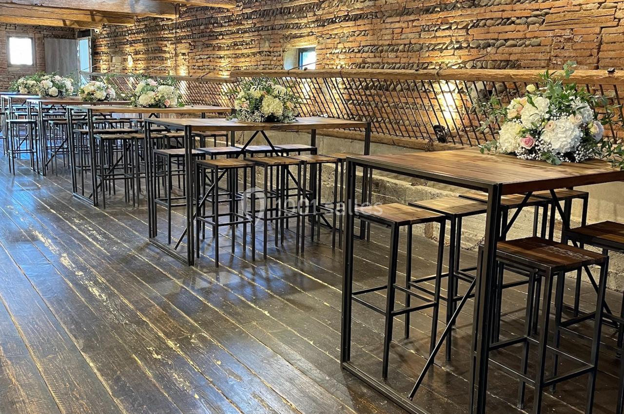 Salle en briques avec poutres apparentes, tables hautes en bois et compositions florales décoratives.