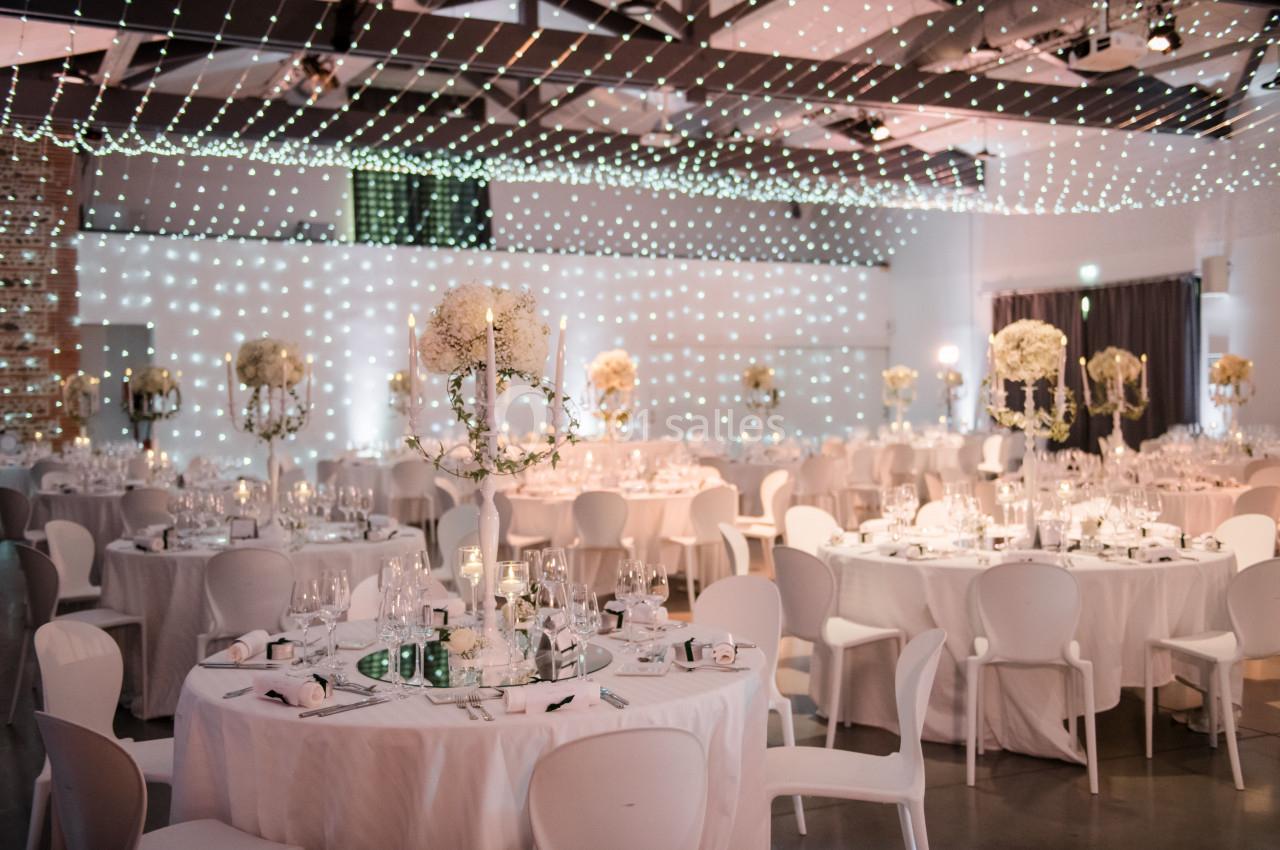 Salle de réception décorée pour un mariage, avec tables rondes, nappes blanches et guirlandes lumineuses au plafond.
