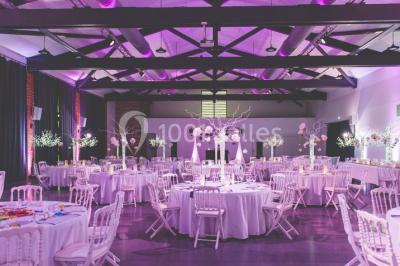 Salle de réception décorée pour un mariage, avec tables rondes, nappes blanches et guirlandes lumineuses au plafond.