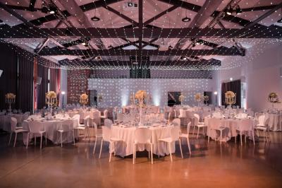 Salle de réception décorée pour un mariage, avec tables rondes, nappes blanches et guirlandes lumineuses au plafond.
