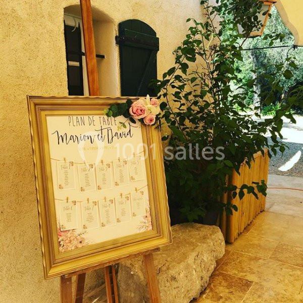 Plan de table encadré avec des fleurs roses, posé sur un chevalet en bois devant un mur clair et une porte verte.