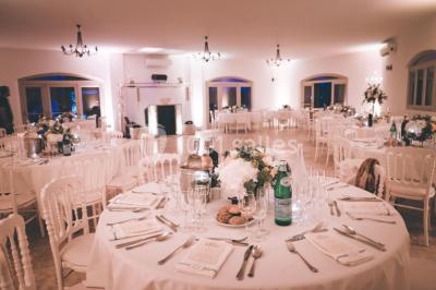 Un couple habillé élégamment, la femme en robe blanche et l'homme en costume, pose dans une pièce lumineuse.