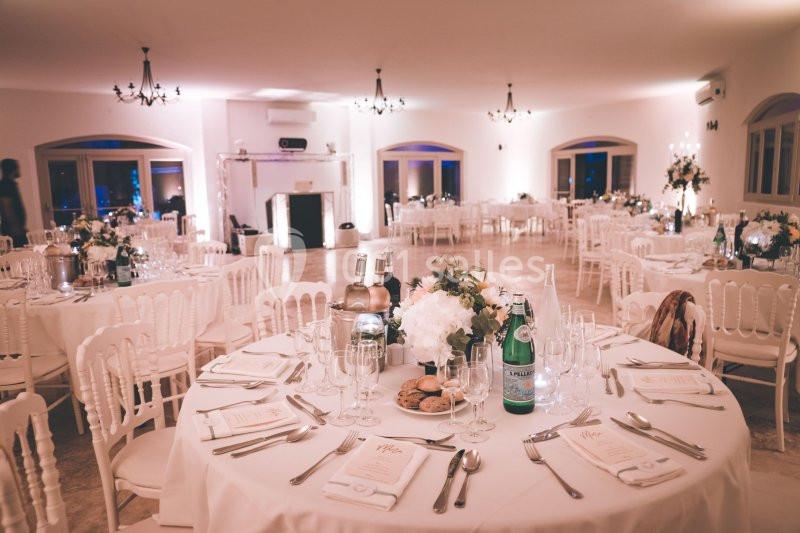 Salle de réception élégamment décorée avec des tables rondes, nappes blanches, vaisselle raffinée et centre de table floral.