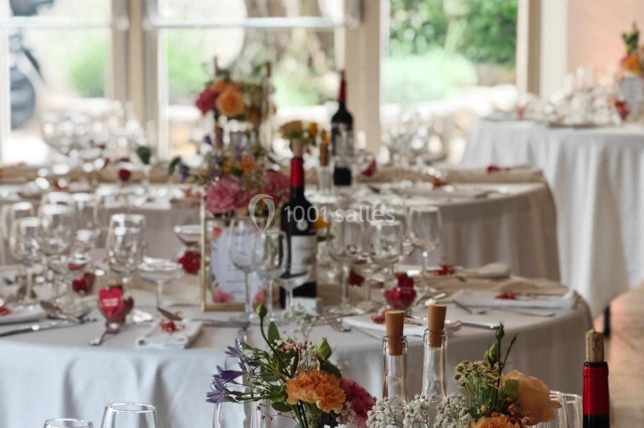 Tables élégamment dressées avec nappes blanches, fleurs colorées, verres et bouteilles de vin dans une salle lumineuse.