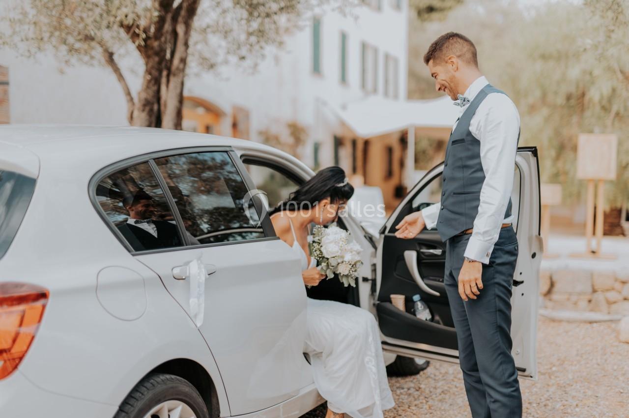 Une femme en robe blanche sort d'une voiture décorée, aidée par un homme en costume, dans un cadre extérieur.