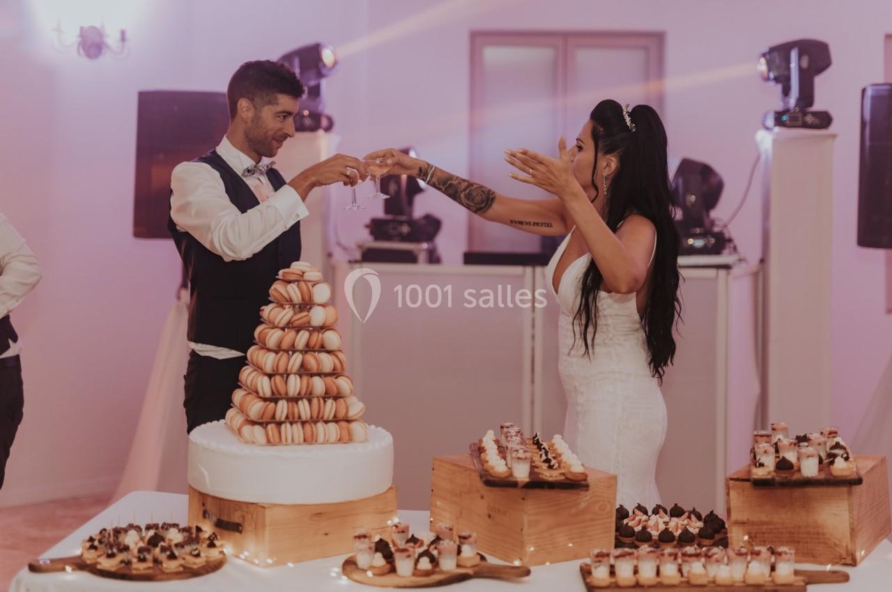 Un couple en tenue de mariage trinque devant une pièce montée de macarons et des desserts sur des tables décorées.