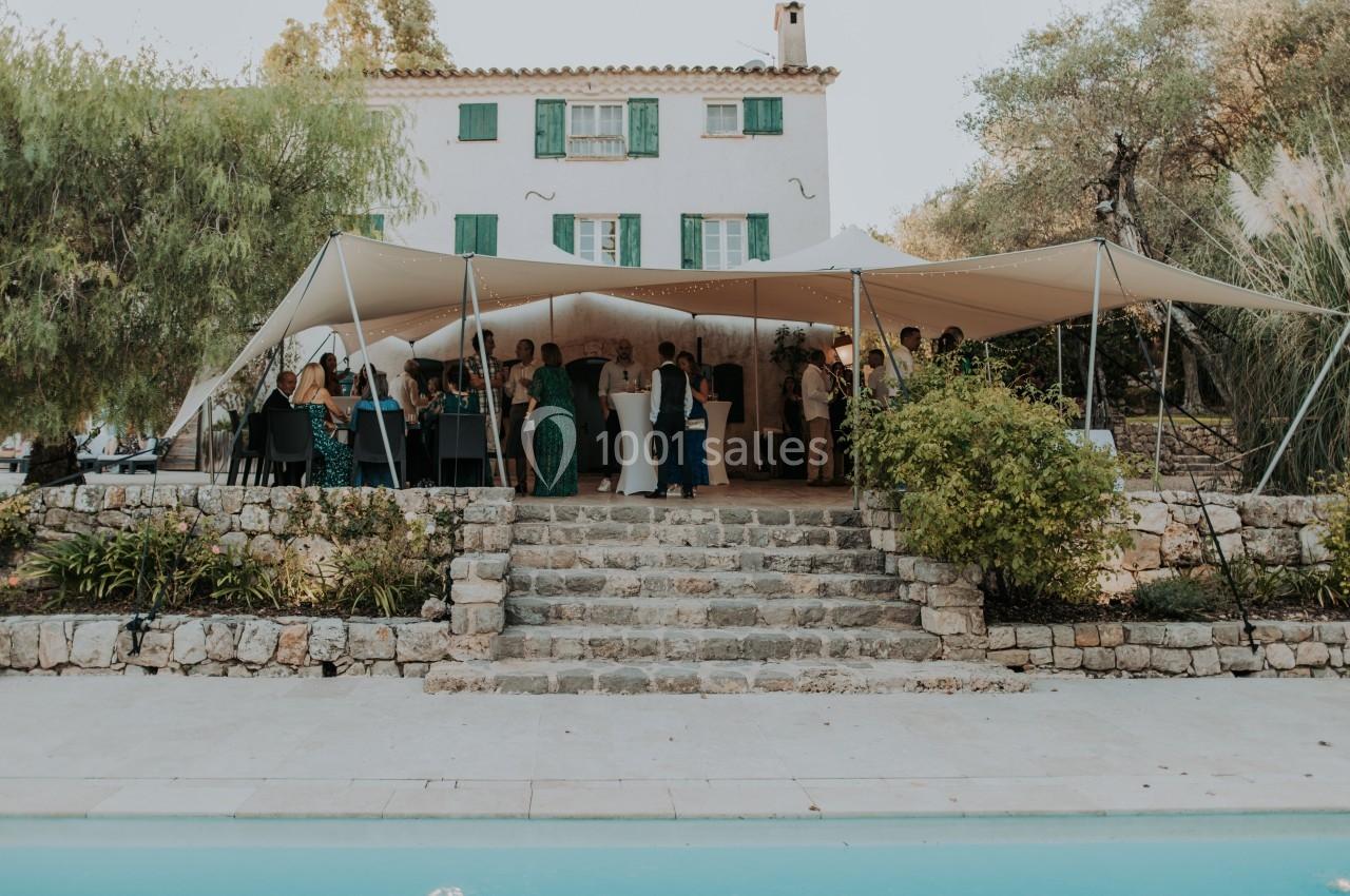 Tente installée dans un jardin devant une maison blanche, avec des invités réunis autour de tables hautes.