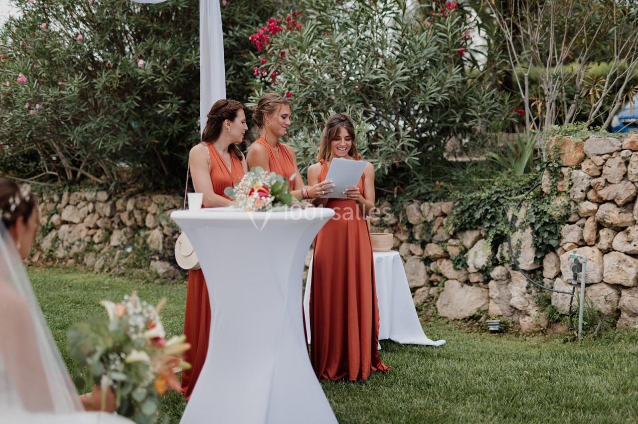 Trois femmes en robes orange lisent un document près d'une table haute lors d'un événement en extérieur.