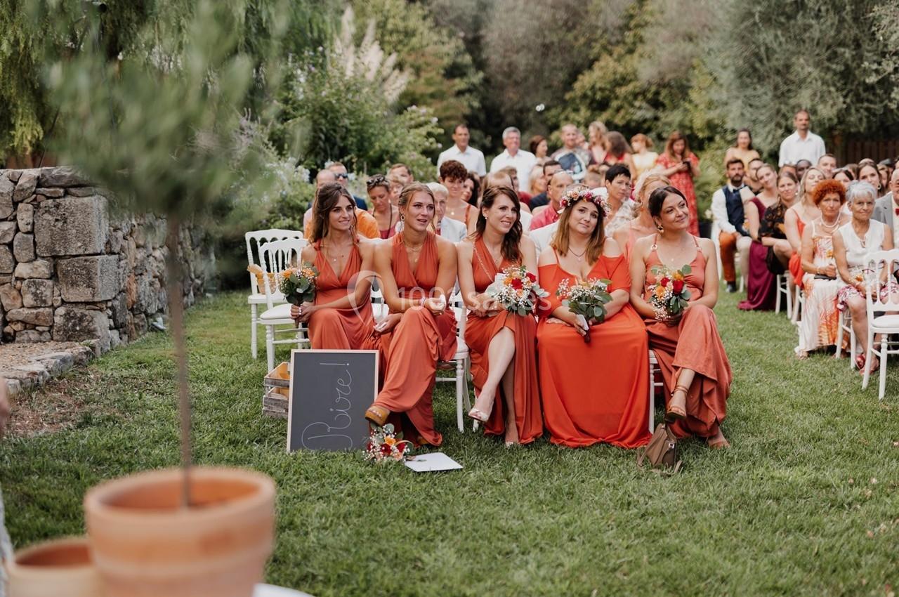 Cinq femmes en robes orange assises sur des chaises dans un jardin, tenant des bouquets, entourées d'invités.