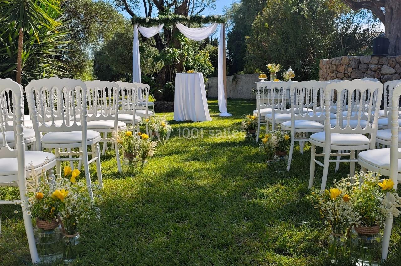 Chaises blanches alignées face à une arche décorée de tissu blanc et fleurs, installées dans un jardin ensoleillé.