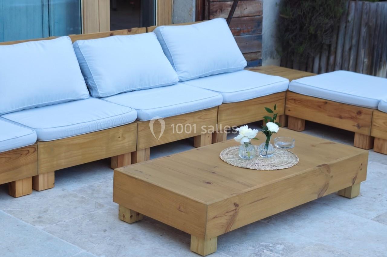 Salon de jardin en bois avec coussins blancs, table basse assortie et décoration florale sur une terrasse en pierre.