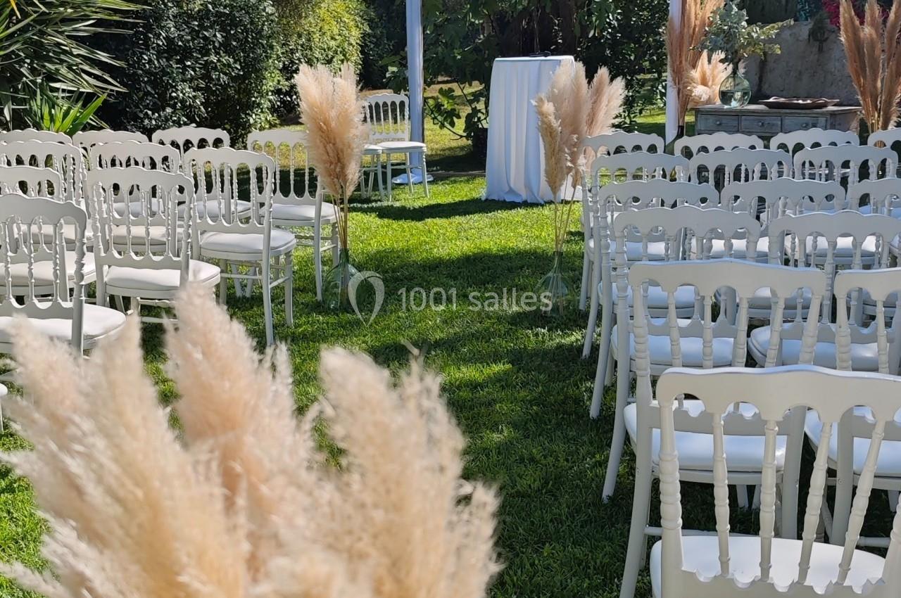 Chaises blanches alignées sur une pelouse entourée de végétation, avec une arche décorée pour une cérémonie en plein air.