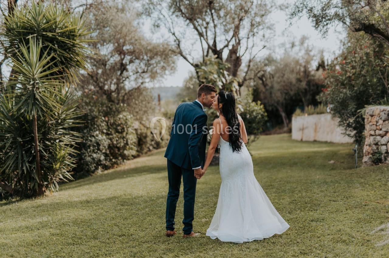 Un couple en tenue de mariage se tient la main et marche dans un jardin verdoyant par une journée ensoleillée.