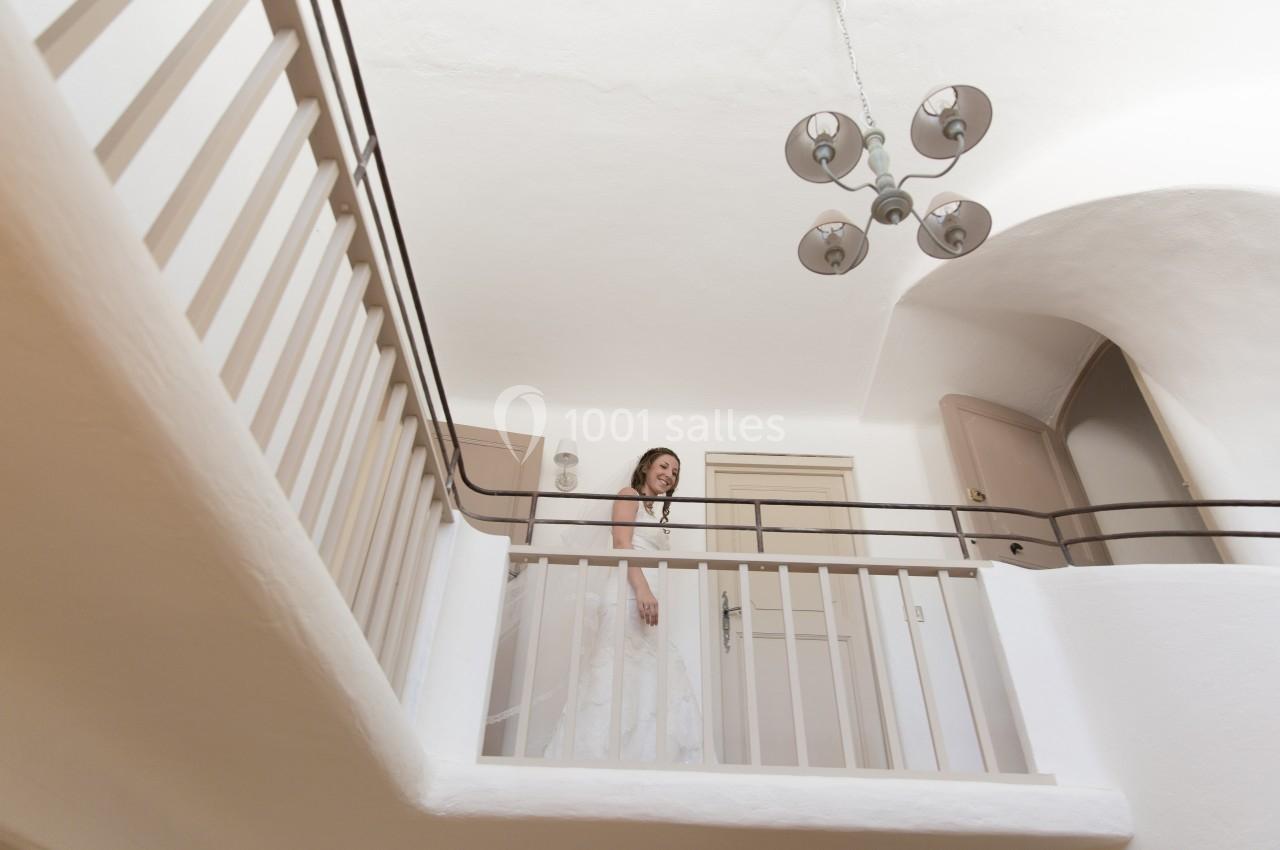 Femme en robe blanche debout sur un balcon intérieur, entourée de murs et plafonds blancs avec une lampe suspendue.