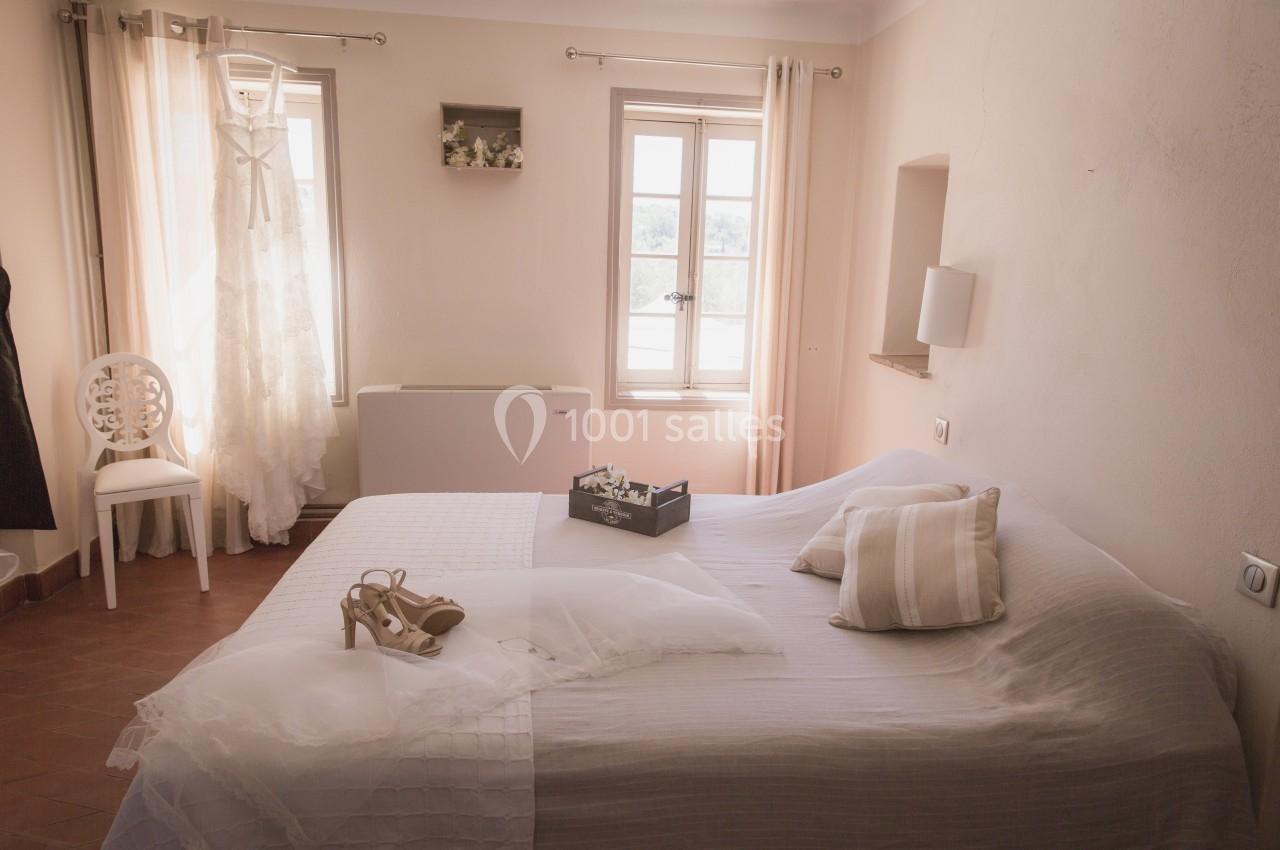 Robe de mariée suspendue près d'une fenêtre dans une chambre lumineuse avec lit décoré d'accessoires de mariage.