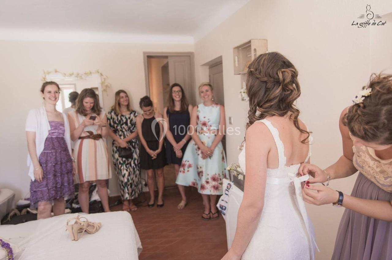 Une mariée se fait ajuster sa robe dans une pièce, entourée de femmes souriantes.