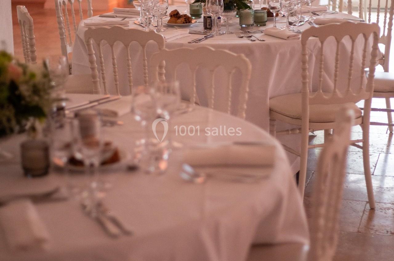 Salle de réception avec des tables rondes dressées, nappes blanches, chaises élégantes et décorations florales.