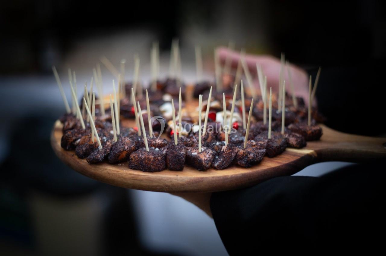 Plateau en bois garni de bouchées grillées avec des pics en bois, tenu par une personne en arrière-plan flou.
